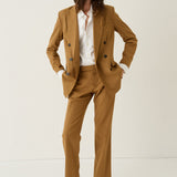 A model wearing a camel brown double-breasted blazer and matching trousers, styled with a white button-down shirt and tan suede shoes, standing against a plain off-white studio background.