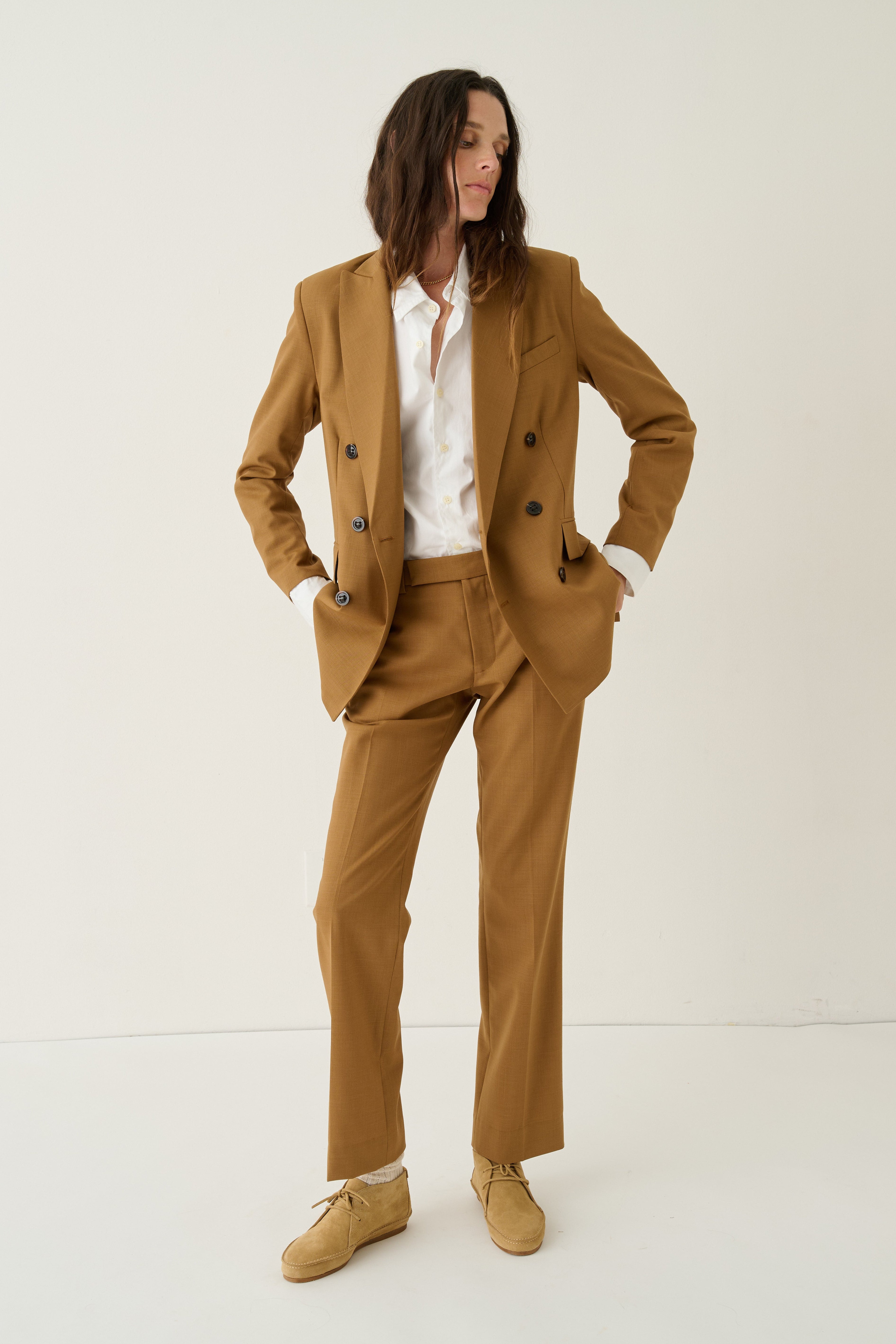 A model wearing a camel brown double-breasted blazer and matching trousers, styled with a white button-down shirt and tan suede shoes, standing against a plain off-white studio background.