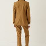 A model viewed from behind wearing a camel brown tailored suit, consisting of a blazer and matching trousers, paired with beige loafers against a plain white studio background.