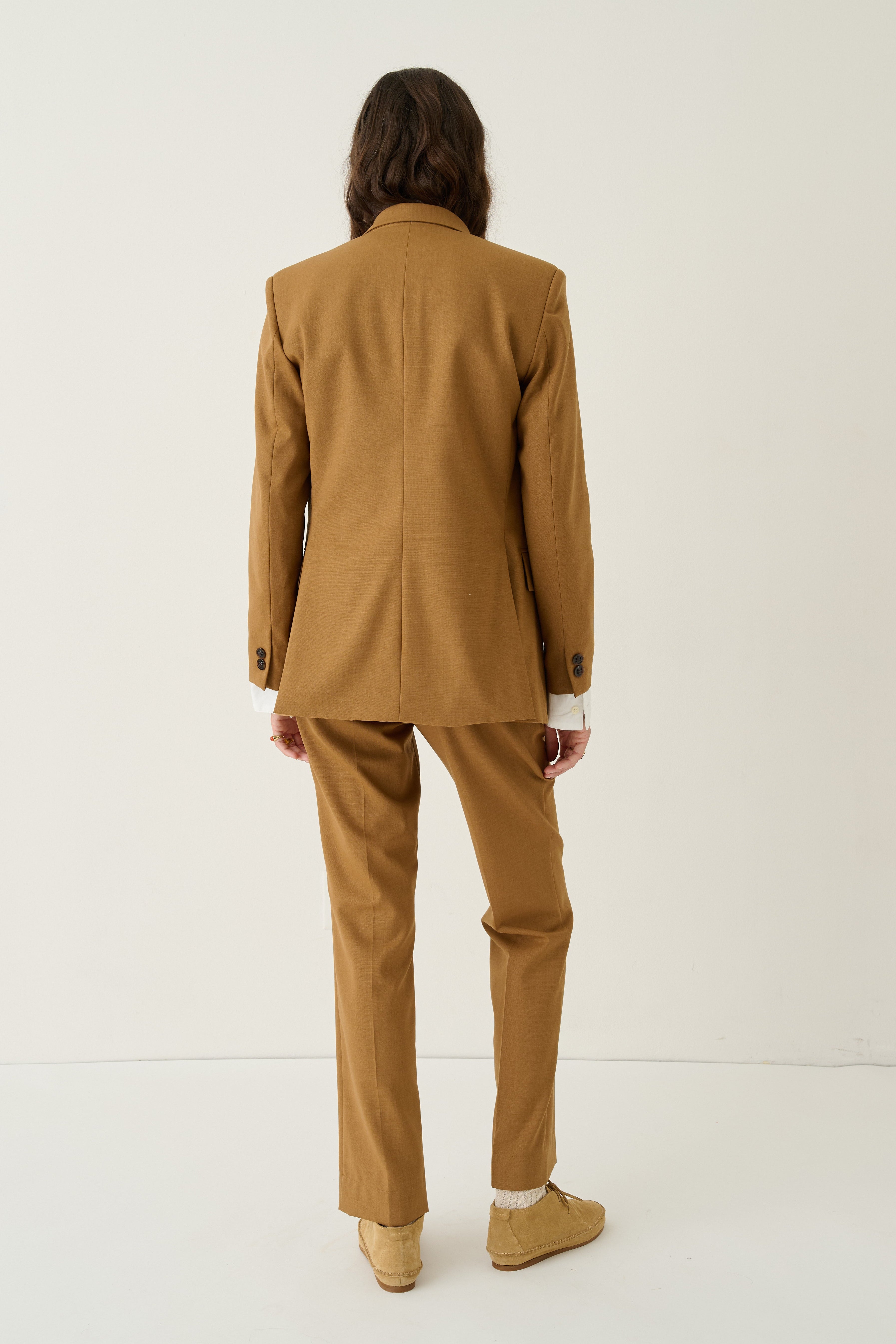 A model viewed from behind wearing a camel brown tailored suit, consisting of a blazer and matching trousers, paired with beige loafers against a plain white studio background.