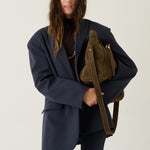 A woman modeling an oversized navy blue blazer and matching trousers, carrying a brown suede shoulder bag with a wide strap against a plain light gray studio background.
