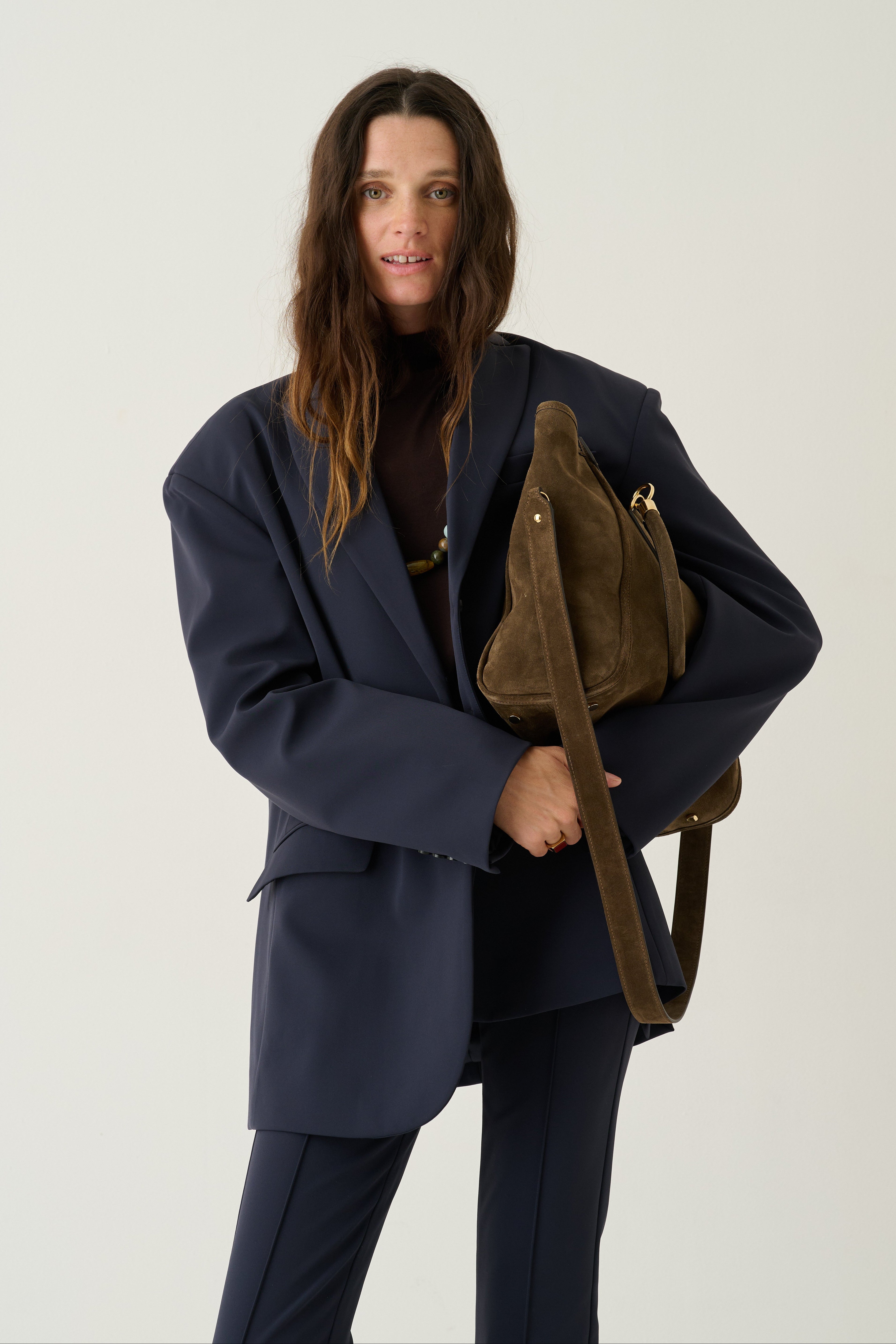 A woman modeling an oversized navy blue blazer and matching trousers, carrying a brown suede shoulder bag with a wide strap against a plain light gray studio background.