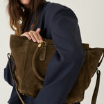 A woman in a navy blue blazer holds a large olive-brown suede tote bag with gold-tone hardware against a plain light background.