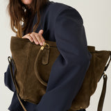 A woman in a navy blue blazer holds a large olive-brown suede tote bag with gold-tone hardware against a plain light background.