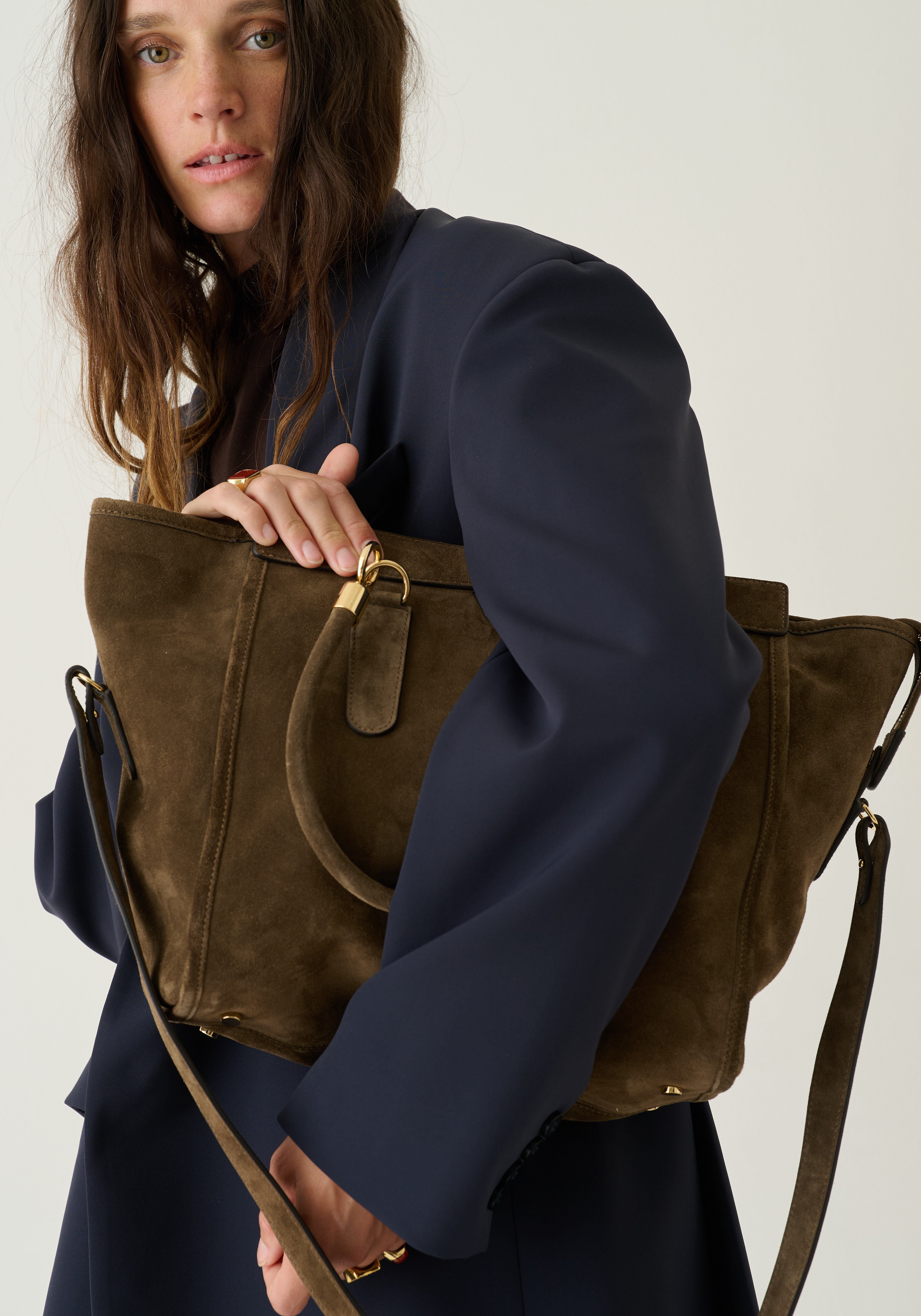 A woman in a navy blue blazer holds a large olive-brown suede tote bag with gold-tone hardware against a plain light background.