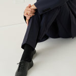 A person sitting on a white floor wearing a dark navy blazer and trousers, black socks, and black lace-up shoes.