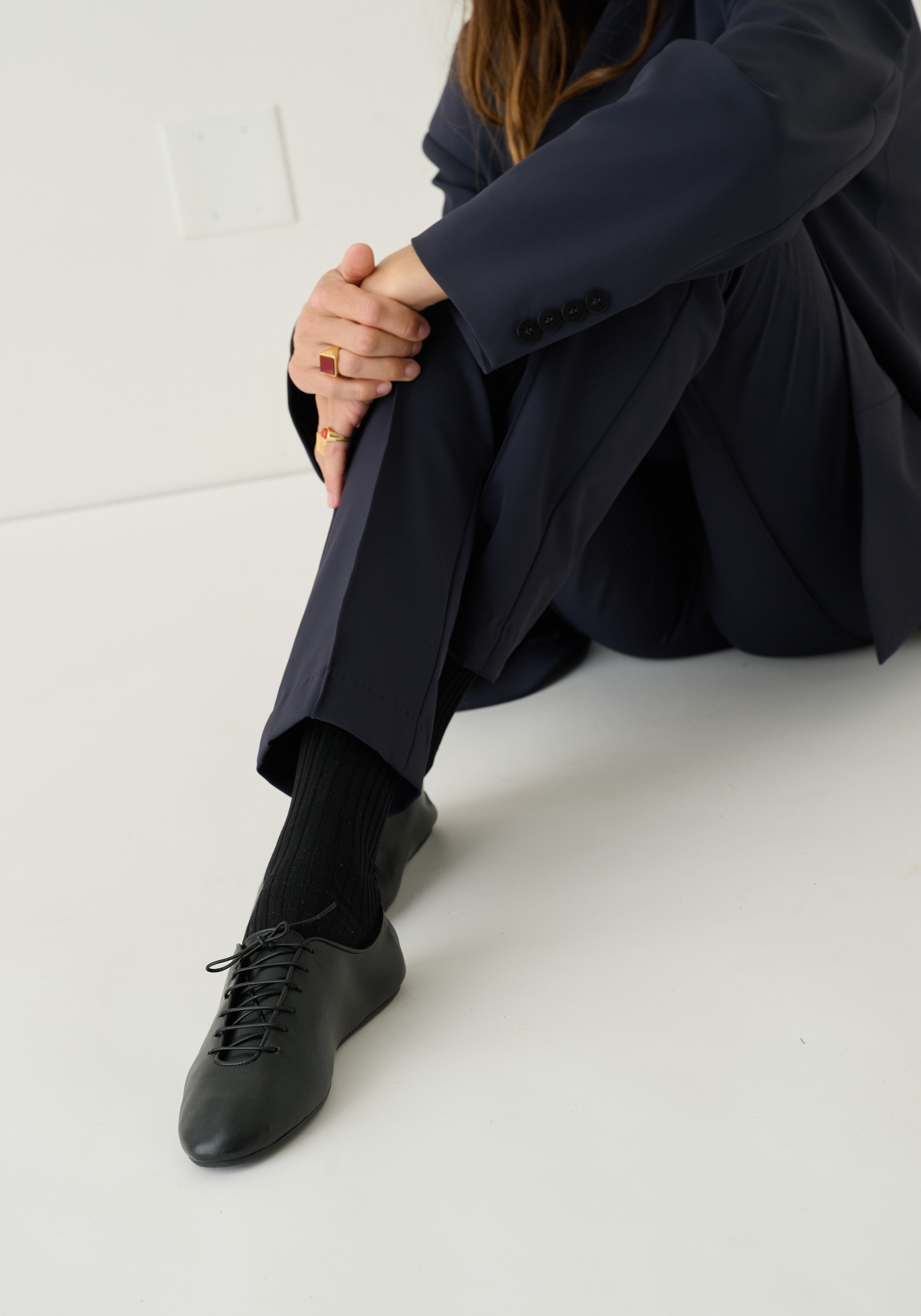 A person sitting on a white floor wearing a dark navy blazer and trousers, black socks, and black lace-up shoes.