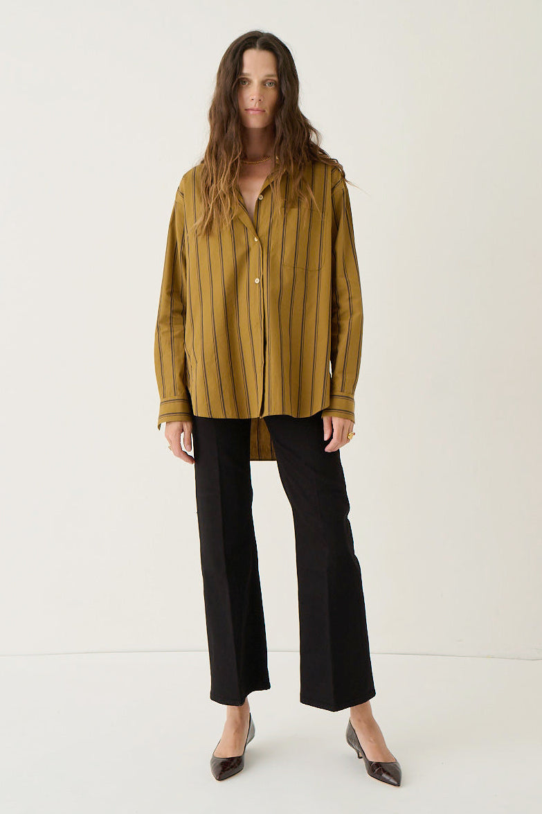 A woman stands against a plain off-white studio background wearing a mustard yellow and black vertically striped button-up shirt, black wide-leg cropped pants, and dark pointed flats.
