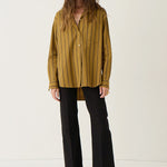 A woman stands against a plain off-white studio background wearing a mustard yellow and black vertically striped button-up shirt, black wide-leg cropped pants, and dark pointed flats.
