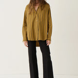 A woman stands against a plain off-white studio background wearing a mustard yellow and black vertically striped button-up shirt, black wide-leg cropped pants, and dark pointed flats.
