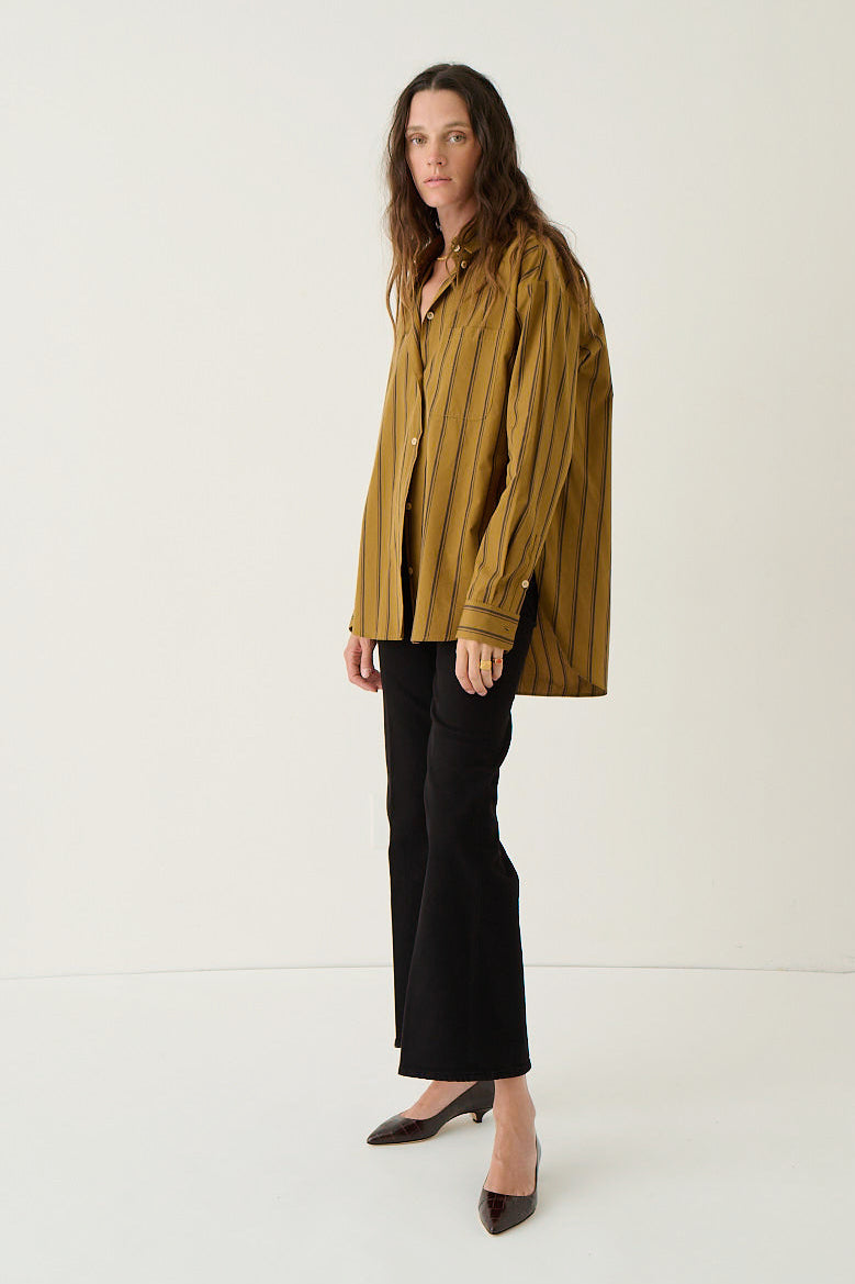 A woman stands in a studio wearing a loose, oversized mustard yellow and black vertically striped button-up shirt, black cropped pants, and dark pointed-toe flats.