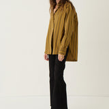 A woman stands in a studio wearing a loose, oversized mustard yellow and black vertically striped button-up shirt, black cropped pants, and dark pointed-toe flats.