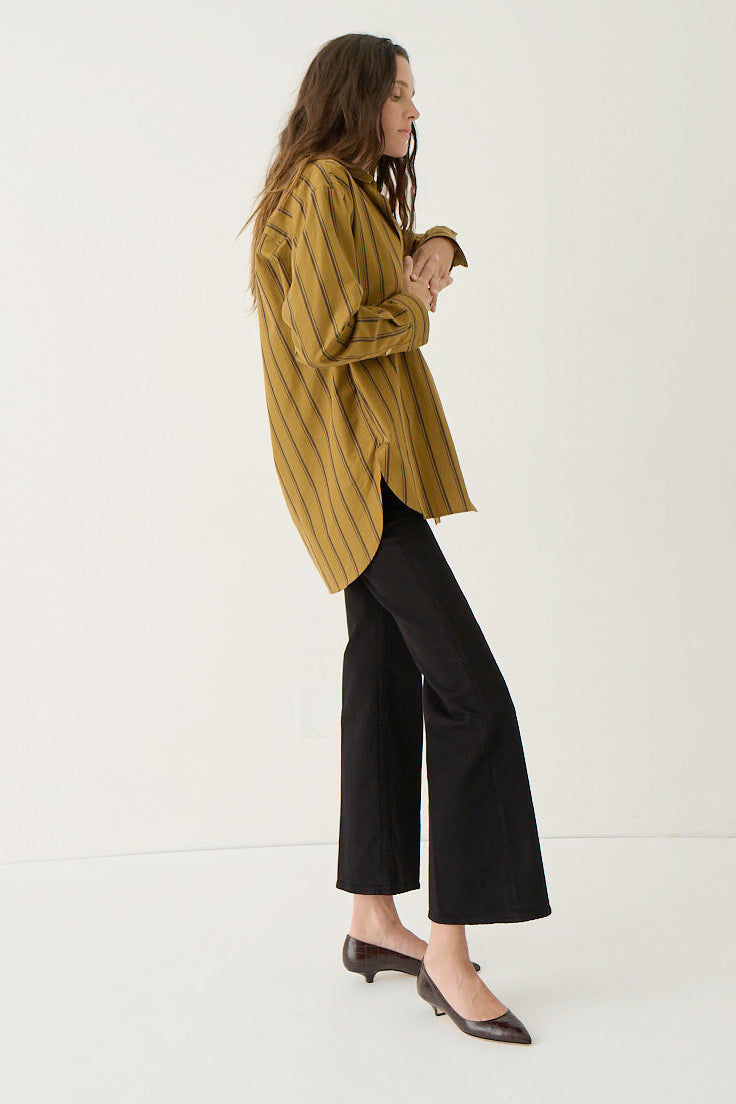 A woman stands in profile wearing an oversized mustard yellow striped button-up shirt, black wide-leg cropped pants, and dark pointed flats against a clean off-white studio background.