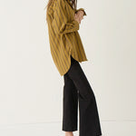 A woman stands in profile wearing an oversized mustard yellow striped button-up shirt, black wide-leg cropped pants, and dark pointed flats against a clean off-white studio background.