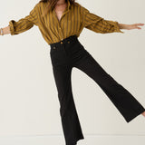 A woman modeling a mustard yellow and black striped long-sleeve button-up shirt paired with black flared pants, posed against a clean off-white studio background.