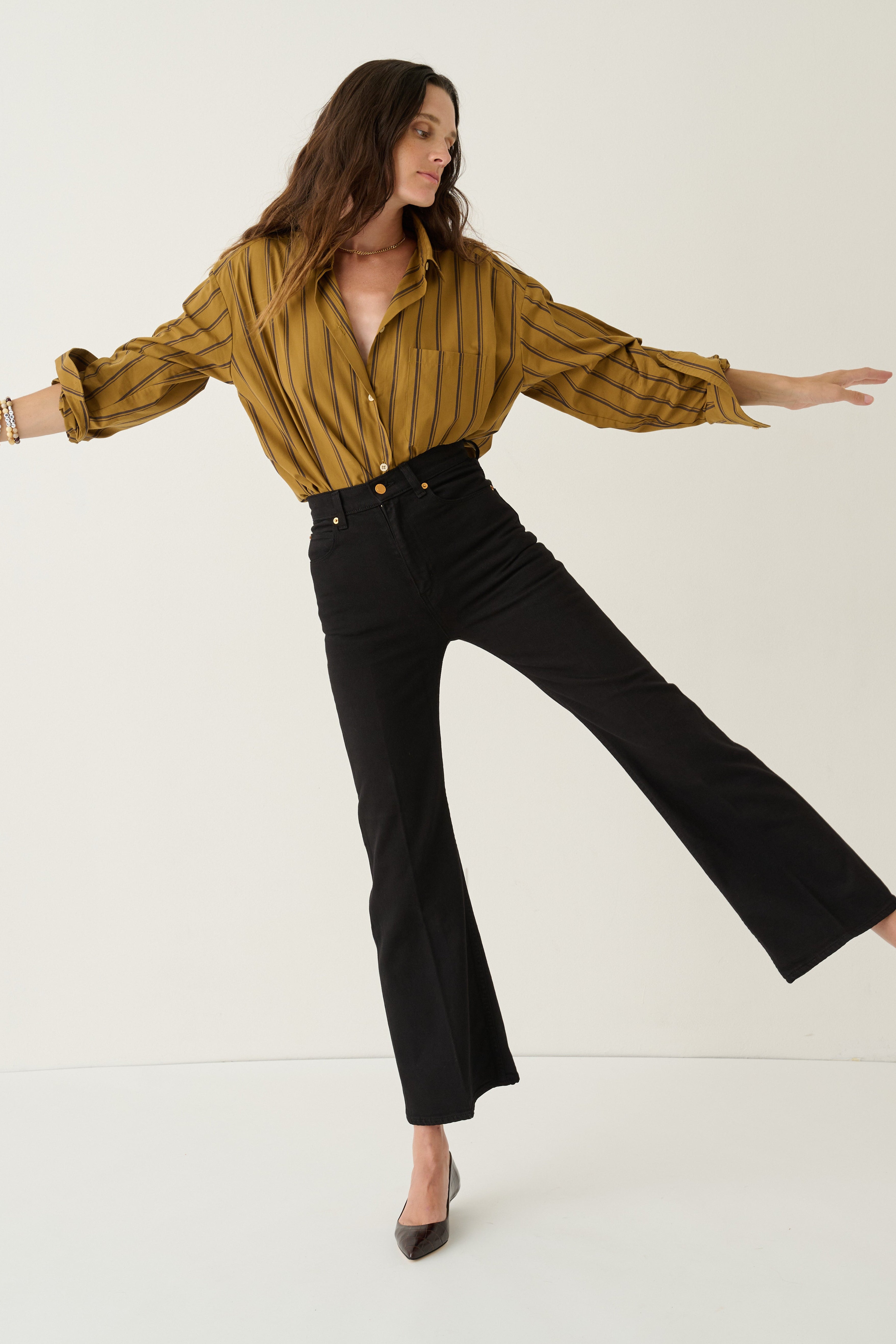 A woman modeling a mustard yellow and black striped long-sleeve button-up shirt paired with black flared pants, posed against a clean off-white studio background.