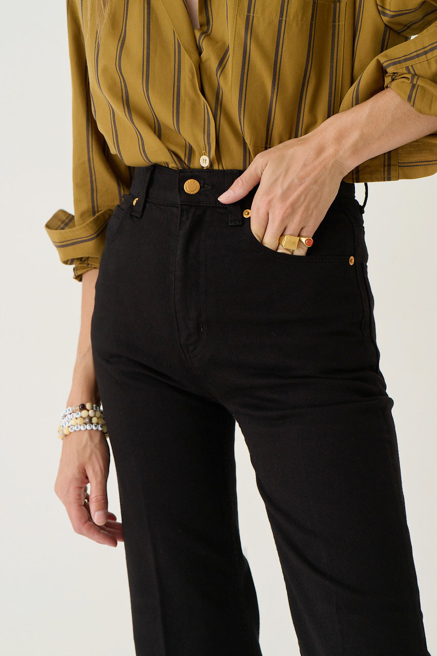 A close-up of a person wearing black high-waisted straight-leg jeans with gold-tone hardware, paired with a mustard yellow striped button-up shirt. The person is shown from the chest down, with one hand in the pocket, against a clean off-white background.