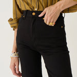 A close-up of a person wearing black high-waisted straight-leg jeans with gold-tone hardware, paired with a mustard yellow striped button-up shirt. The person is shown from the chest down, with one hand in the pocket, against a clean off-white background.