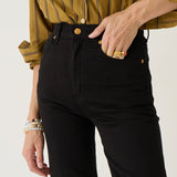 A close-up of a person wearing black high-waisted straight-leg jeans with gold-tone hardware, paired with a mustard yellow striped button-up shirt. The person is shown from the chest down, with one hand in the pocket, against a clean off-white background.