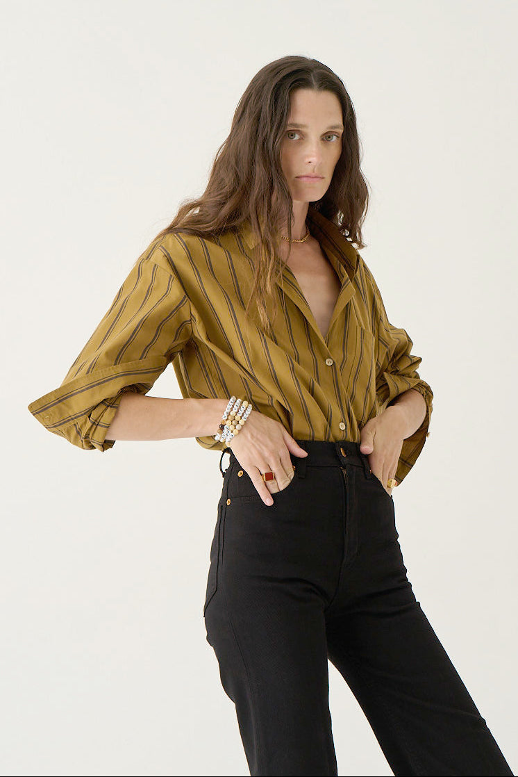 A woman modeling a mustard yellow button-down shirt with thin vertical black stripes, paired with black pants, against a plain white studio background.