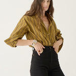 A woman modeling a mustard yellow button-down shirt with thin vertical black stripes, paired with black pants, against a plain white studio background.