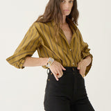 A woman modeling a mustard yellow button-down shirt with thin vertical black stripes, paired with black pants, against a plain white studio background.
