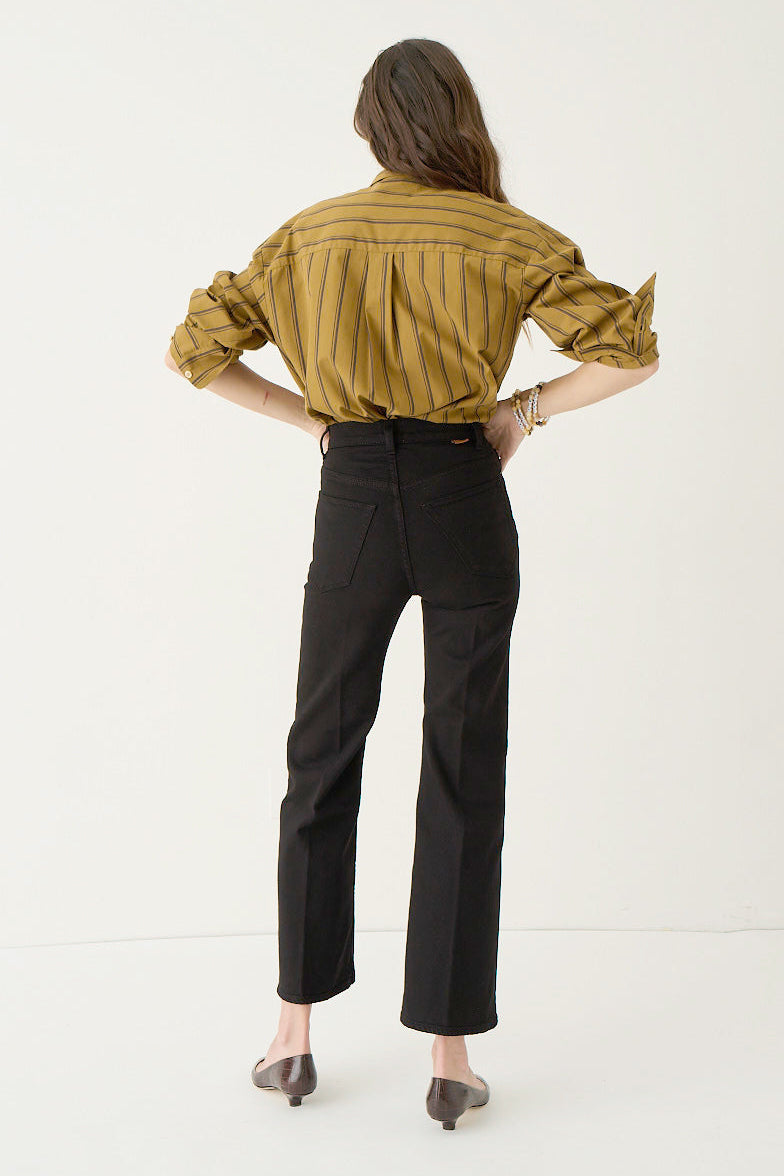 A woman shown from the back wearing a mustard yellow and black vertically striped button-down shirt, black cropped trousers, and dark pointed flats, standing against a plain white studio background.