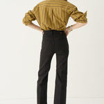 A woman shown from the back wearing a mustard yellow and black vertically striped button-down shirt, black cropped trousers, and dark pointed flats, standing against a plain white studio background.