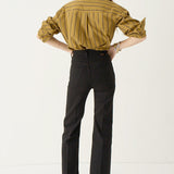 A woman shown from the back wearing a mustard yellow and black vertically striped button-down shirt, black cropped trousers, and dark pointed flats, standing against a plain white studio background.