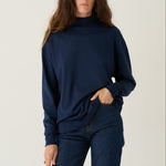 A woman modeling a navy blue long-sleeve mock neck sweater and matching navy blue jeans against a plain light gray studio background.