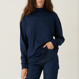 A woman modeling a navy blue long-sleeve mock neck sweater and matching navy blue jeans against a plain light gray studio background.