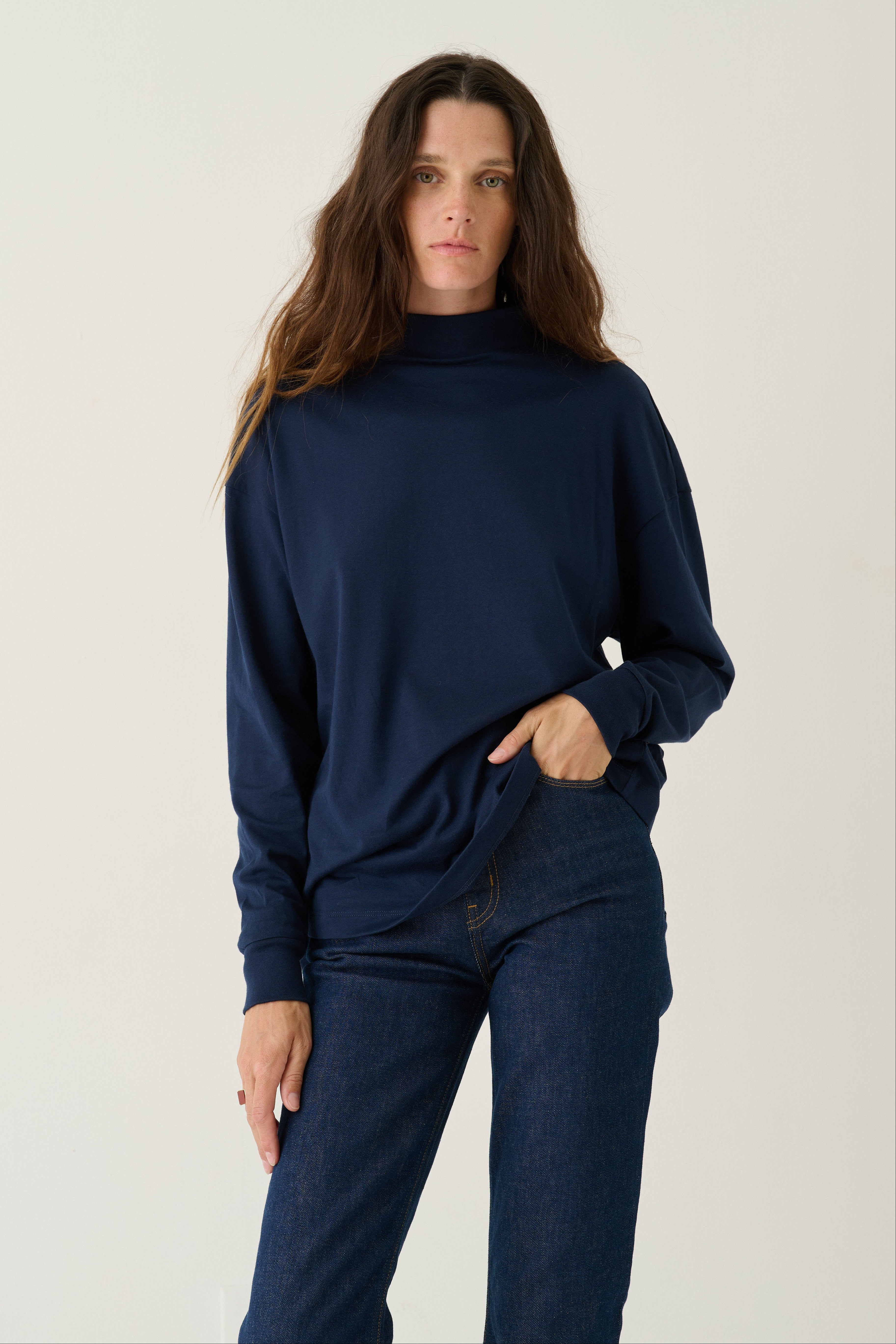 A woman modeling a navy blue long-sleeve mock neck sweater and matching navy blue jeans against a plain light gray studio background.