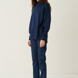 A woman stands in profile wearing a dark navy blue mock-neck long-sleeve top and matching navy blue pants, paired with black shoes, against a plain off-white studio background.