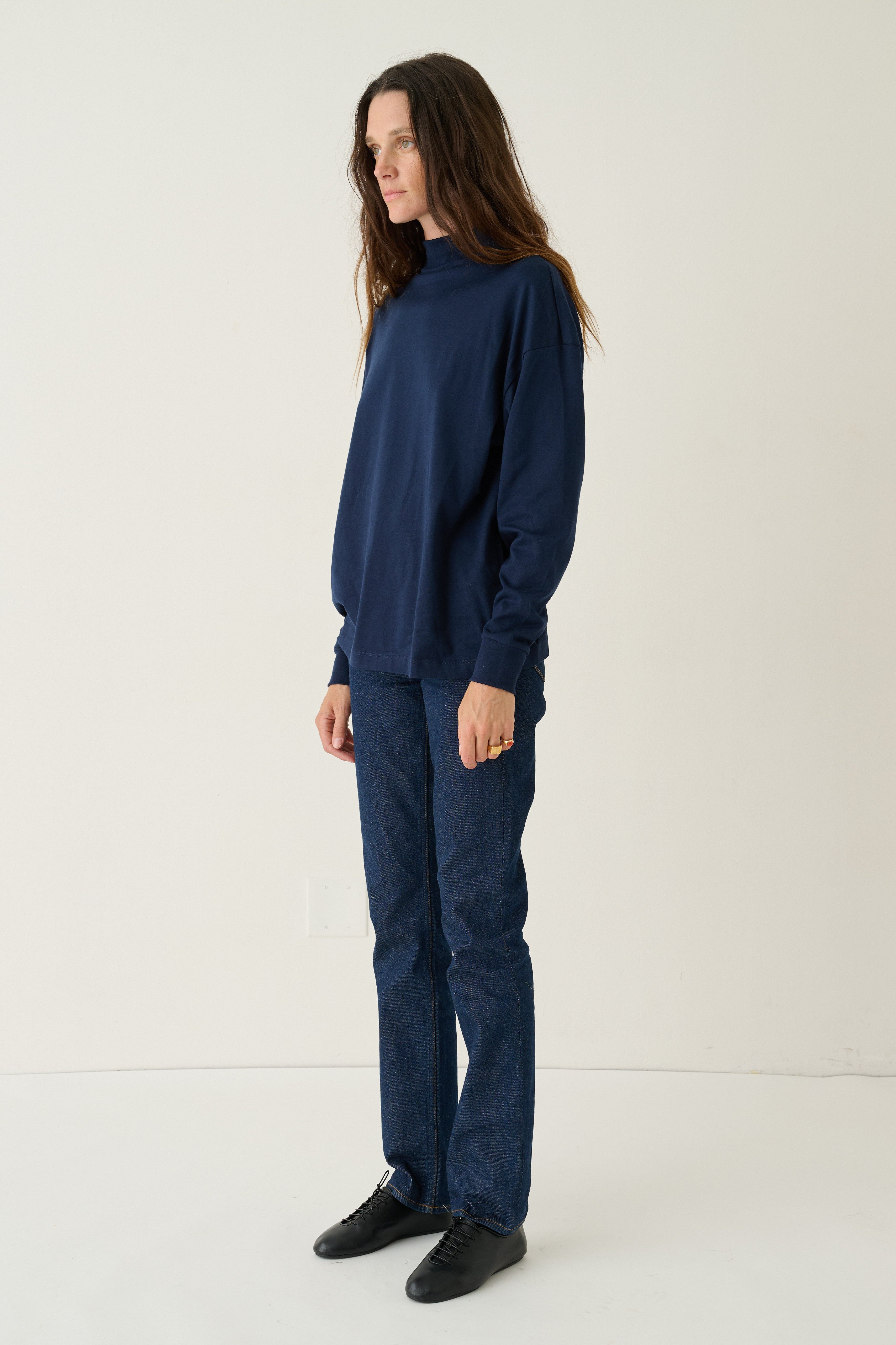 A woman stands in profile wearing a dark navy blue mock-neck long-sleeve top and matching navy blue pants, paired with black shoes, against a plain off-white studio background.