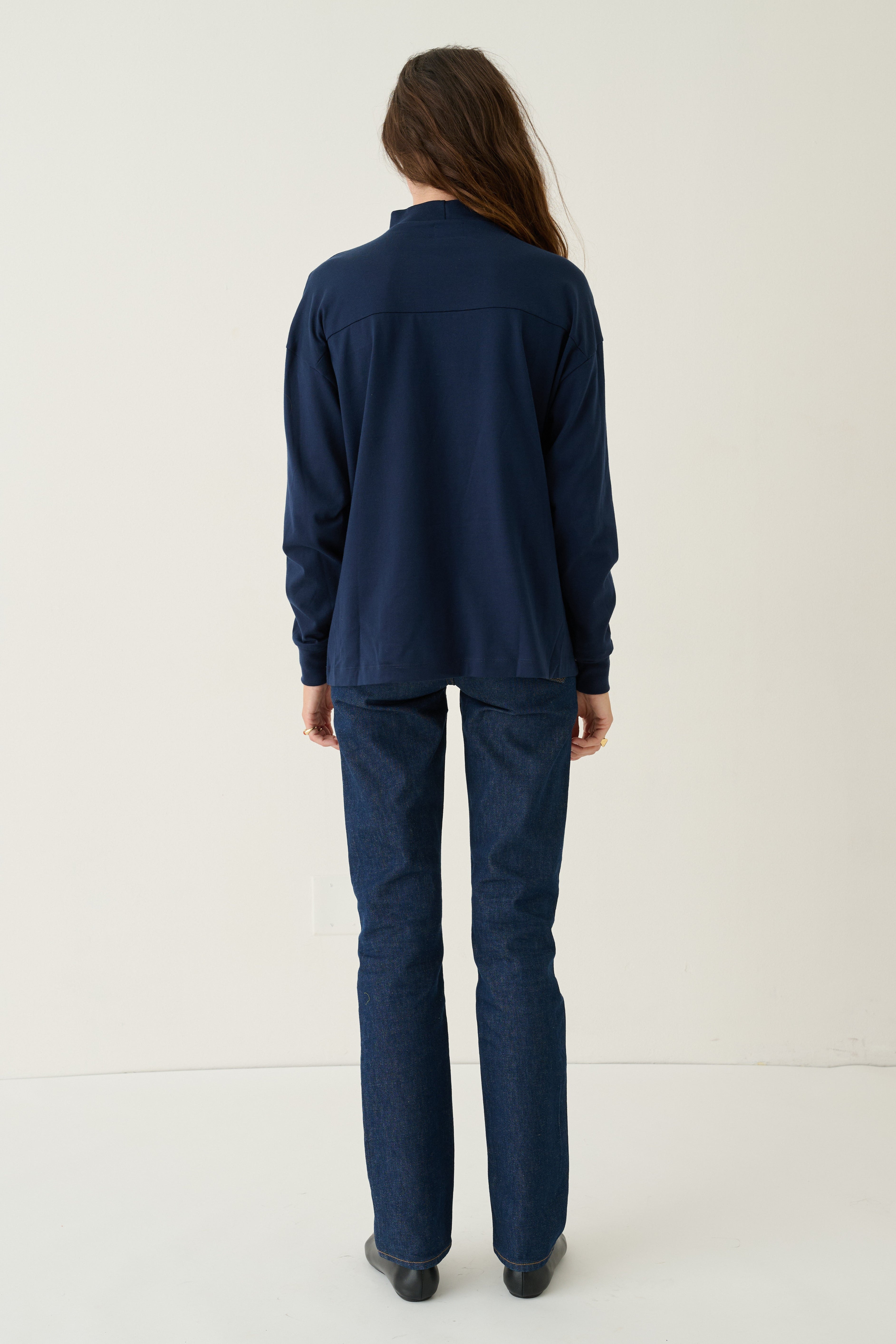 A woman viewed from behind wearing a matching navy blue long-sleeve top and trousers set, standing against a plain off-white studio background.