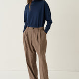 A model stands against a plain off-white studio background wearing a navy blue mock-neck long-sleeve top, brown high-waisted pleated trousers, and black shoes.