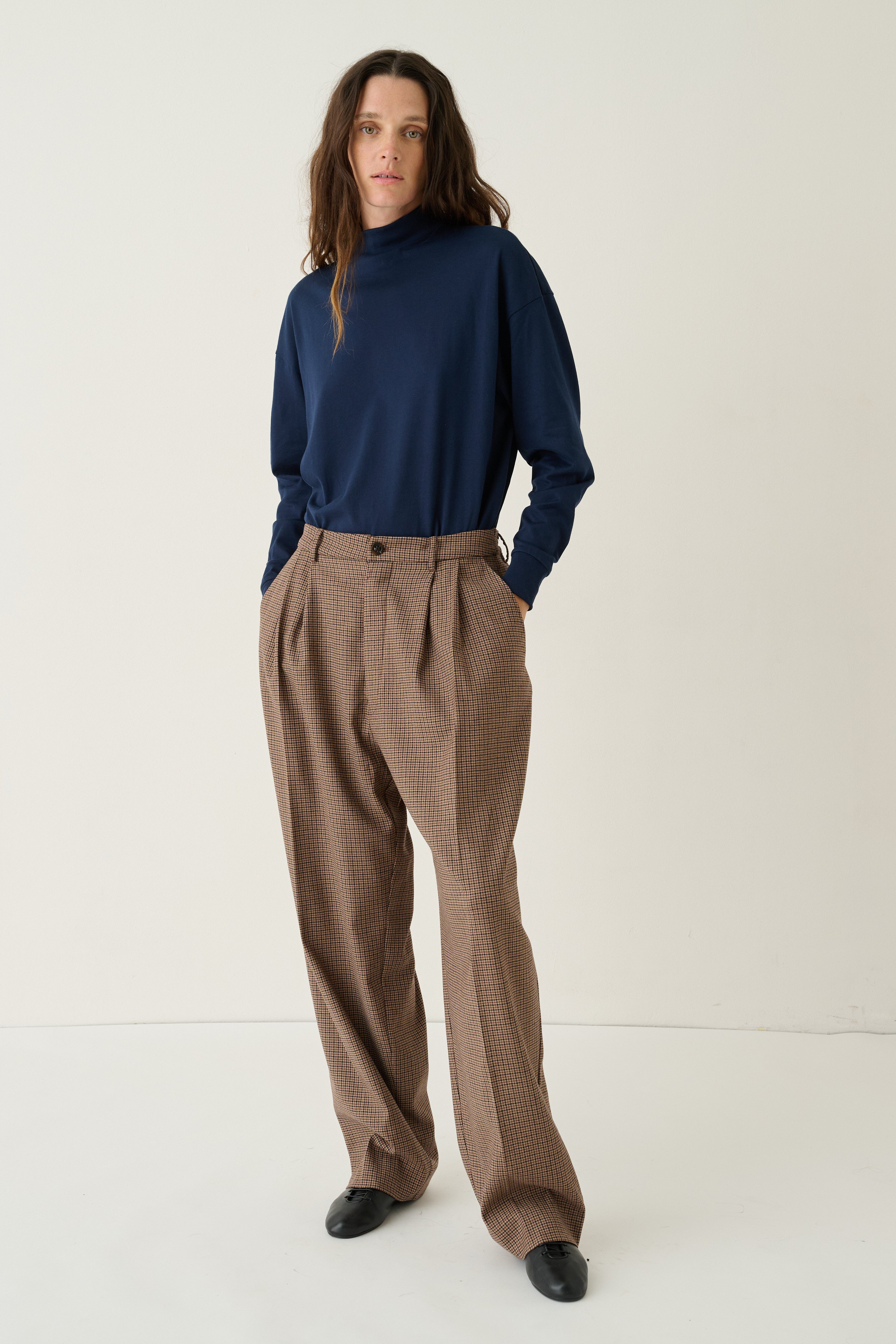 A model stands against a plain off-white studio background wearing a navy blue mock-neck long-sleeve top, brown high-waisted pleated trousers, and black shoes.
