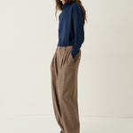 A woman stands in a studio wearing a navy blue long-sleeve top and loose brown wide-leg trousers, paired with black shoes, against a plain off-white background.