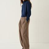 A woman stands in a studio wearing a navy blue long-sleeve top and loose brown wide-leg trousers, paired with black shoes, against a plain off-white background.