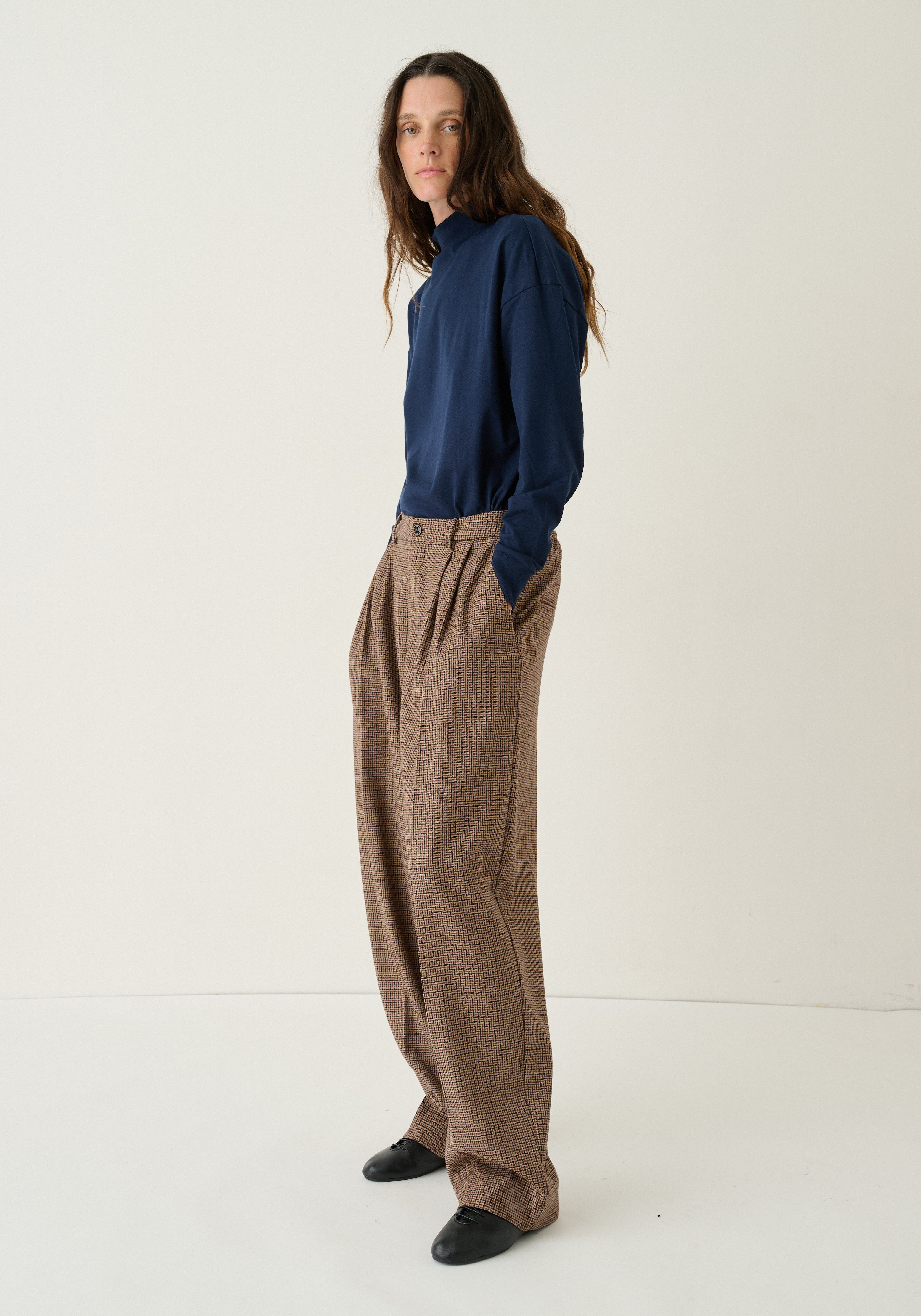 A woman stands in a studio wearing a navy blue long-sleeve top and loose brown wide-leg trousers, paired with black shoes, against a plain off-white background.