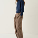 A woman stands in a studio wearing a navy blue long-sleeve top and loose brown wide-leg trousers, paired with black shoes, against a plain off-white background.