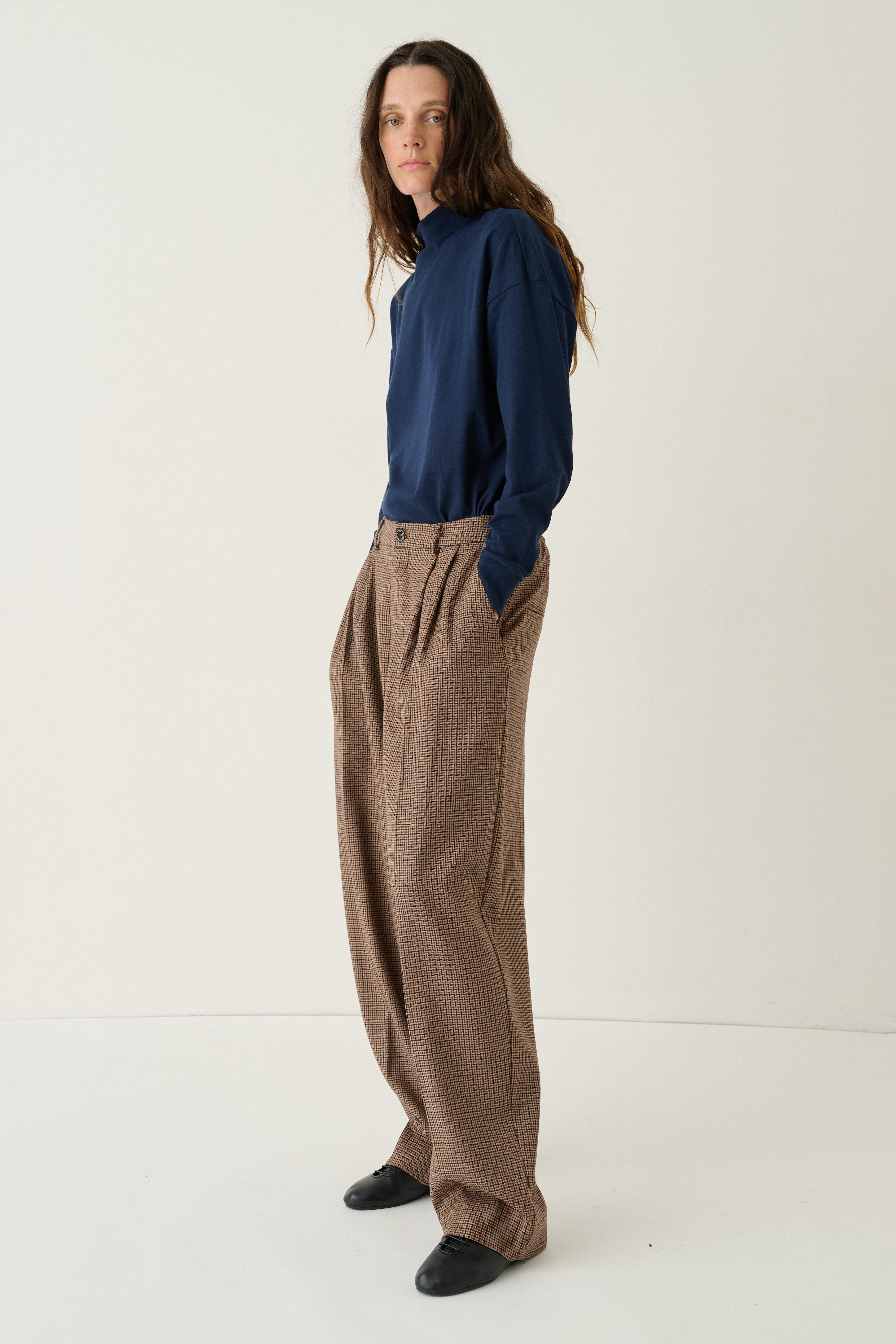 A woman stands in a studio wearing a navy blue long-sleeve top and loose brown wide-leg trousers, paired with black shoes, against a plain off-white background.
