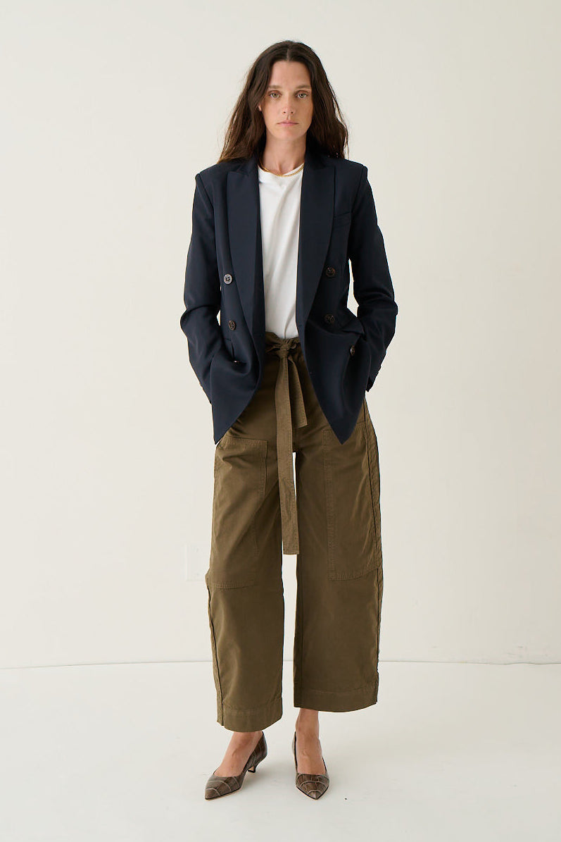 A woman stands against a plain off-white studio background wearing a dark navy blazer over a white top, paired with olive green wide-leg cropped pants and brown pointed-toe flats.