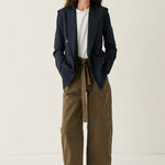 A woman stands against a plain off-white studio background wearing a dark navy blazer over a white top, paired with olive green wide-leg cropped pants and brown pointed-toe flats.
