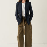 A woman stands against a plain off-white studio background wearing a dark navy blazer over a white top, paired with olive green wide-leg cropped pants and brown pointed-toe flats.