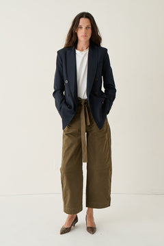 A woman stands against a plain off-white studio background wearing a dark navy blazer over a white top, paired with olive green wide-leg cropped pants and brown pointed-toe flats.