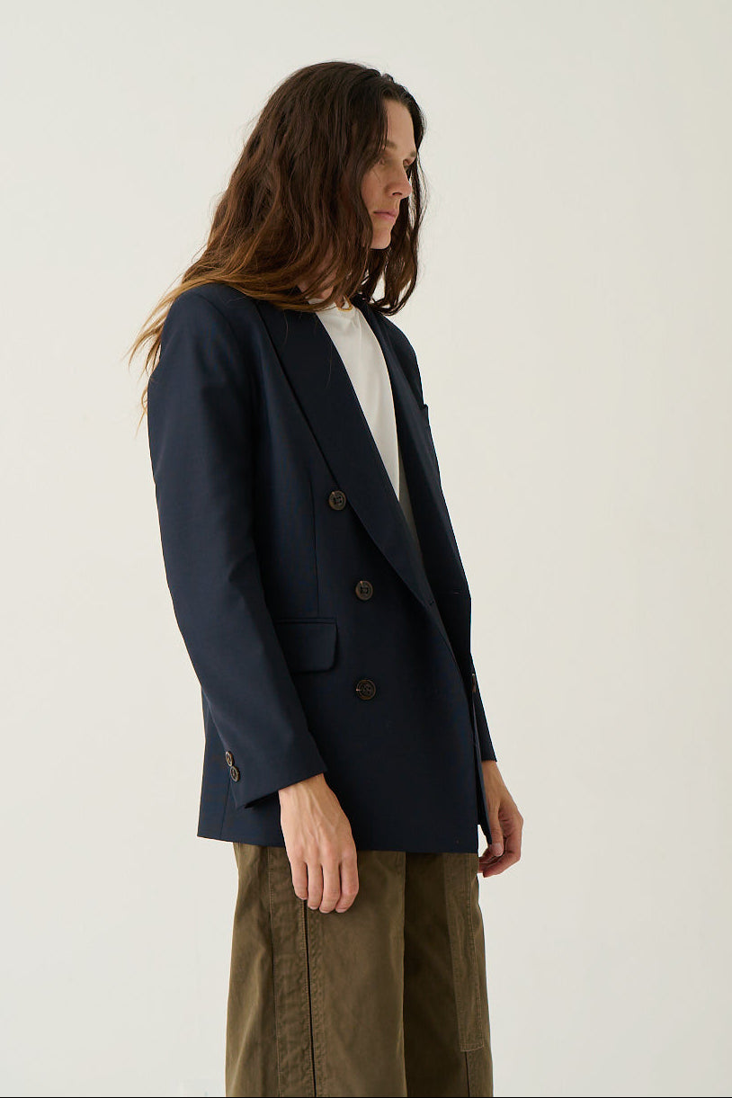A model wearing a navy blue double-breasted blazer over a white top and olive green trousers, standing against a plain off-white studio background.