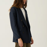 A model wearing a navy blue double-breasted blazer over a white top and olive green trousers, standing against a plain off-white studio background.