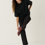 A woman modeling a black oversized short-sleeve top and black wide-leg trousers, paired with dark brown leather ankle boots, against a plain off-white studio background.