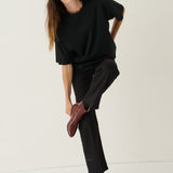 A woman modeling a black oversized short-sleeve top and black wide-leg trousers, paired with dark brown leather ankle boots, against a plain off-white studio background.